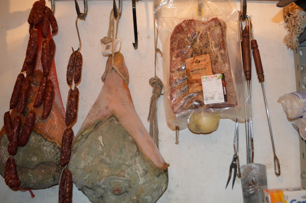 Various types of cured meats, including sausages, hams, and a packaged pancetta, are hanging from hooks. The meats are arranged on a wall, showing a rustic and artisanal display. Cooking tools with wooden handles are also hanging next to the meats.
