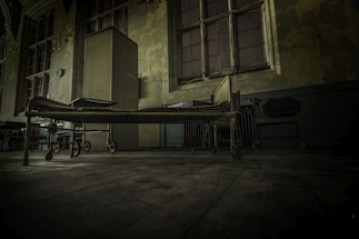 An old, abandoned room with peeling wallpaper and a worn-out hospital bed on wheels. The wooden floor is dusty, and the windows are large with cracked glass panes, allowing minimal light into the dimly lit space. A tall, metallic cabinet and some old furniture add to the desolate ambiance.