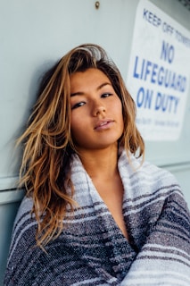 woman with black, white, and grey blanket leaning on wall with no lifeguard on duty signage