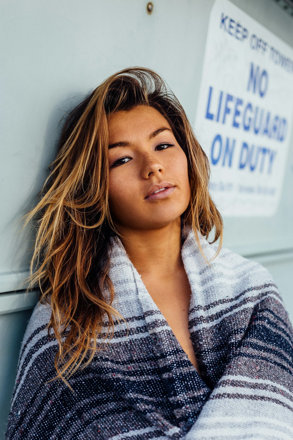 woman with black, white, and grey blanket leaning on wall with no lifeguard on duty signage