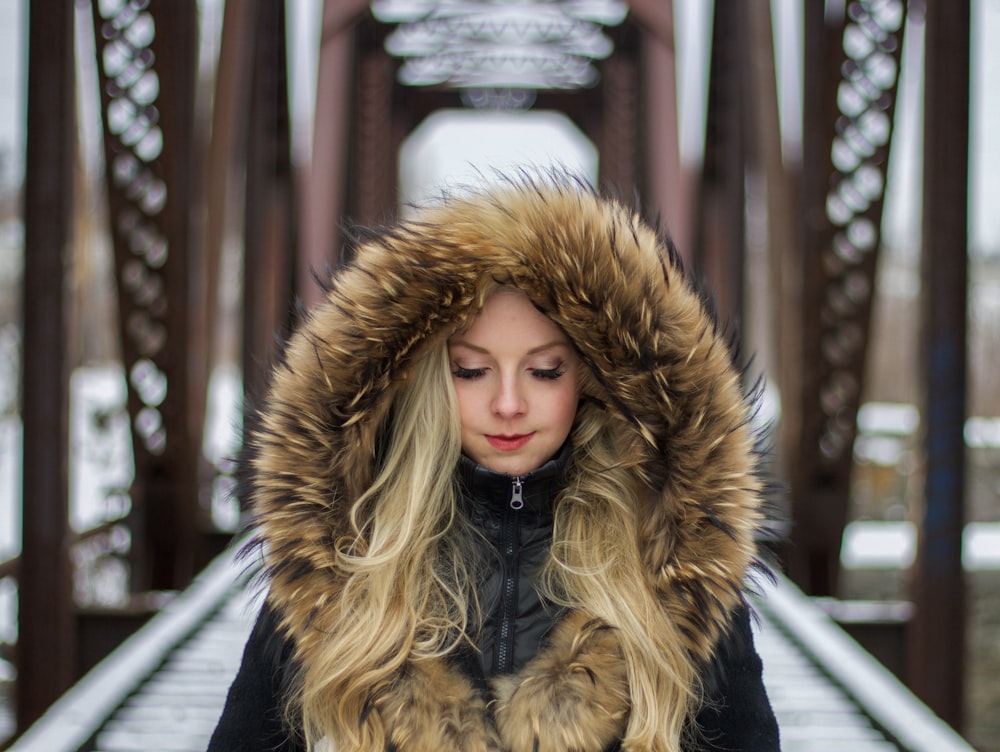 Donna che indossa una felpa con cappuccio foderata di pelliccia marrone in piedi sul ponte durante la fotografia diurna