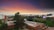 A university campus with modern buildings is set against a vibrant, colorful sky during sunset. The landscape includes lush green lawns, trees, and a central building with a prominent sign. In the background, a large digital screen displays an image.