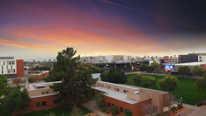 A university campus with modern buildings is set against a vibrant, colorful sky during sunset. The landscape includes lush green lawns, trees, and a central building with a prominent sign. In the background, a large digital screen displays an image.