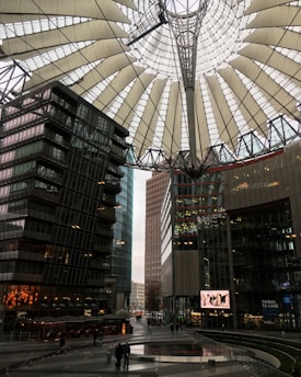 A modern urban architecture scene featuring a large, distinctive canopy above a public square. Surrounding the square are several tall, glass-covered buildings reflecting the daylight. There are people walking around, and a digital screen displays content. The setting appears to be a sophisticated, bustling city environment.