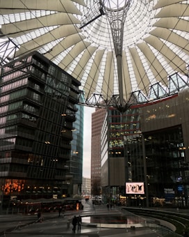 A modern urban architecture scene featuring a large, distinctive canopy above a public square. Surrounding the square are several tall, glass-covered buildings reflecting the daylight. There are people walking around, and a digital screen displays content. The setting appears to be a sophisticated, bustling city environment.