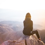 person sitting on top of gray rock overlooking mountain during daytime