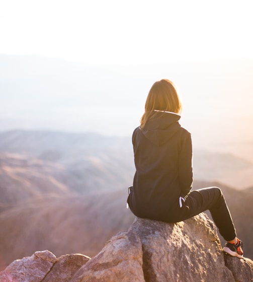 person sitting on top of gray rock overlooking mountain during daytime