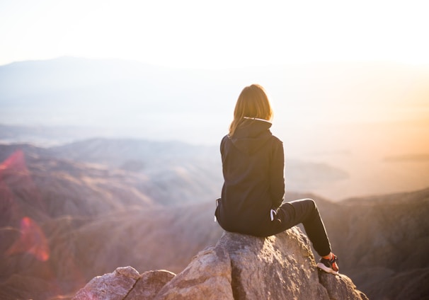 person sitting on top of gray rock overlooking mountain during daytime