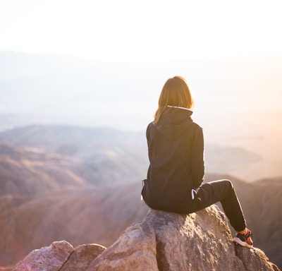 person sitting on top of gray rock overlooking mountain during daytime