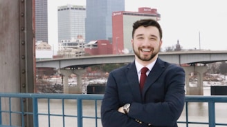 A person wearing a suit and tie is standing in front of an urban landscape with tall buildings and a river. They are smiling and appear to be confident and relaxed.