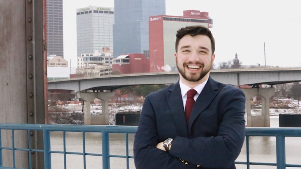 A person wearing a suit and tie is standing in front of an urban landscape with tall buildings and a river. They are smiling and appear to be confident and relaxed.