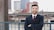 A person wearing a suit and tie is standing in front of an urban landscape with tall buildings and a river. They are smiling and appear to be confident and relaxed.