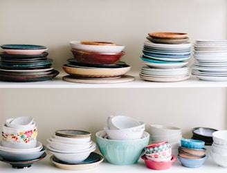 A collection of stacked ceramic bowls and plates of various colors and styles arranged neatly on two white shelves. The dishes include a variety of sizes, with some featuring floral patterns or solid colors, ranging from muted pastels to darker tones.
