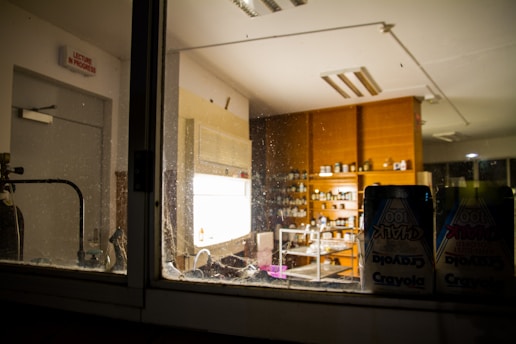 A view of a dimly lit laboratory through a glass window, showcasing shelves filled with various bottles and containers. The room contains wooden cabinets and a sink. A visible chalkboard sign on the wall states 'Lecture in Progress,' and packs of Crayola chalk are in the foreground.