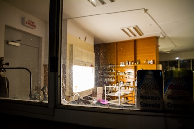 A view of a dimly lit laboratory through a glass window, showcasing shelves filled with various bottles and containers. The room contains wooden cabinets and a sink. A visible chalkboard sign on the wall states 'Lecture in Progress,' and packs of Crayola chalk are in the foreground.