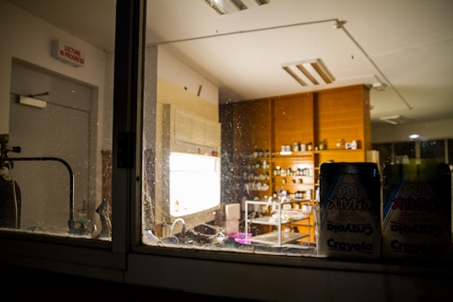A view of a dimly lit laboratory through a glass window, showcasing shelves filled with various bottles and containers. The room contains wooden cabinets and a sink. A visible chalkboard sign on the wall states 'Lecture in Progress,' and packs of Crayola chalk are in the foreground.