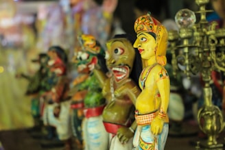 Colorful traditional figurines, crafted with intricate details, standing in a row. The figures display a vibrant array of primary colors and are arranged alongside a decorative, ornate metal object resembling a candelabra.