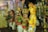 Colorful traditional figurines, crafted with intricate details, standing in a row. The figures display a vibrant array of primary colors and are arranged alongside a decorative, ornate metal object resembling a candelabra.
