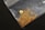 A small pile of gold flakes and a larger gold nugget are placed on a transparent plastic bag against a dark background. The gold pieces vary in size and shape, with the nugget being distinctly larger and rounder.