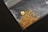 A small pile of gold flakes and a larger gold nugget are placed on a transparent plastic bag against a dark background. The gold pieces vary in size and shape, with the nugget being distinctly larger and rounder.