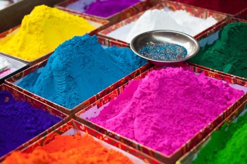 Various piles of vibrant colored powders are neatly arranged in square sections. The colors include bright blue, pink, yellow, green, orange, purple, and white. A small metallic bowl containing colorful glitter is placed on top of one of the sections, adding to the array of colors.