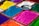 Various piles of vibrant colored powders are neatly arranged in square sections. The colors include bright blue, pink, yellow, green, orange, purple, and white. A small metallic bowl containing colorful glitter is placed on top of one of the sections, adding to the array of colors.