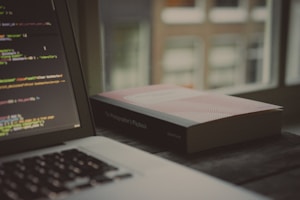 An open laptop displaying a code editor with lines of code is situated on a wooden surface. Next to it lies a closed book titled 'The Photographer's Playbook'. The backdrop consists of blurred windows suggesting an indoor setting with soft, natural light filtering through.