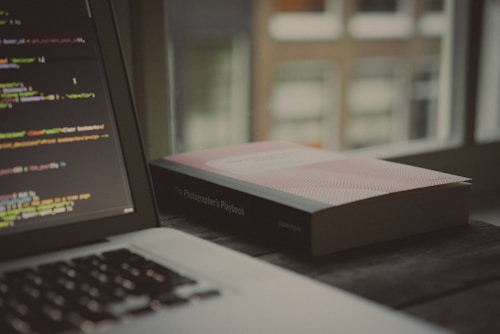 An open laptop displaying a code editor with lines of code is situated on a wooden surface. Next to it lies a closed book titled 'The Photographer's Playbook'. The backdrop consists of blurred windows suggesting an indoor setting with soft, natural light filtering through.