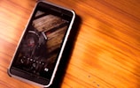 A smartphone with a rugged black and white case is placed on a wooden surface. The screen displays a close-up image of a camera with visible details of the lens and the Canon logo.