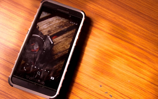 A smartphone with a rugged black and white case is placed on a wooden surface. The screen displays a close-up image of a camera with visible details of the lens and the Canon logo.