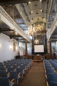 A large room with a high, ornate ceiling featuring decorative chandeliers. The room is arranged like a conference or lecture hall with rows of empty blue-grey chairs facing a stage. There is a projection screen on the stage, flanked by decorative wooden columns and intricate wall designs. The room has a historic and grand ambiance, with detailed moldings and artworks visible on the walls.