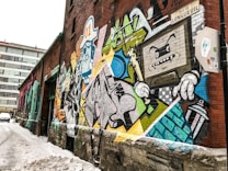 A vibrant street art mural painted on a brick wall along a snowy alleyway. The mural displays a colorful array of graffiti featuring cartoon-like characters, abstract shapes, and a variety of bold lines. A prominent detail includes a TV-headed character with jagged teeth, holding a spray can.