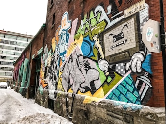 A vibrant street art mural painted on a brick wall along a snowy alleyway. The mural displays a colorful array of graffiti featuring cartoon-like characters, abstract shapes, and a variety of bold lines. A prominent detail includes a TV-headed character with jagged teeth, holding a spray can.
