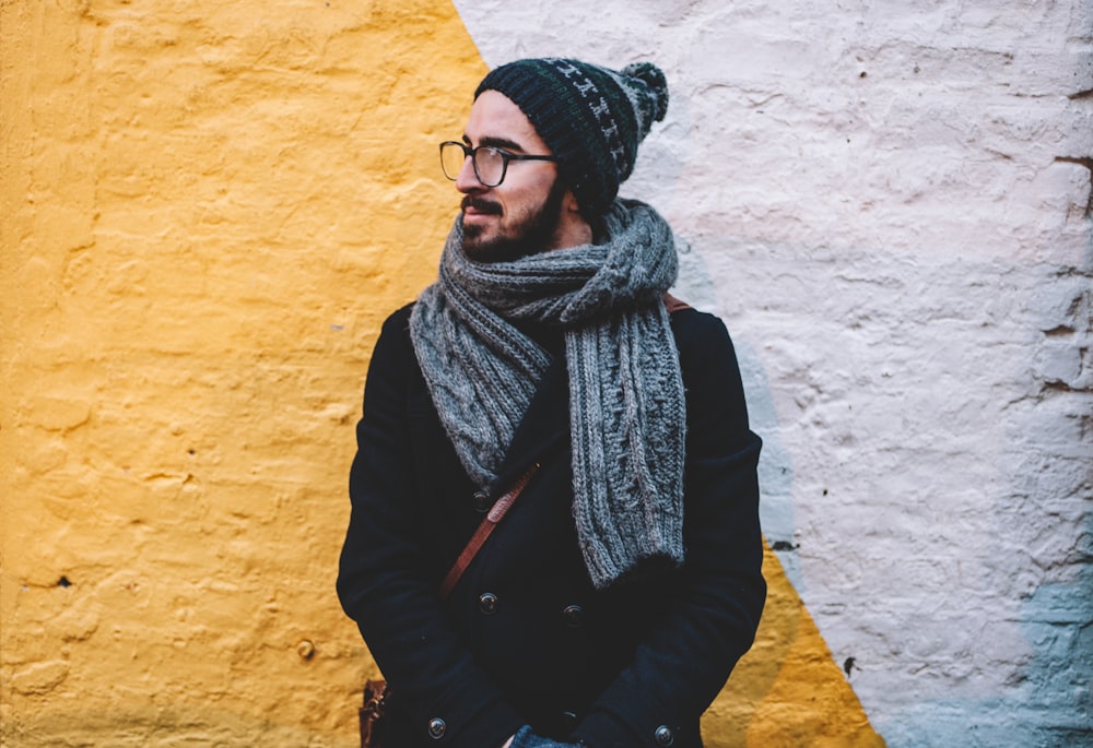 man in black coat with grey scarf standing on front of white and yellow painted wall