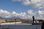 A person is standing on the edge of a rooftop parking area, holding a camera to take photographs. The sky is filled with fluffy white clouds, and in the background, there are red cranes of a port and a distant view of a city skyline with mountains.