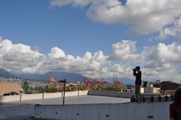 A person is standing on the edge of a rooftop parking area, holding a camera to take photographs. The sky is filled with fluffy white clouds, and in the background, there are red cranes of a port and a distant view of a city skyline with mountains.