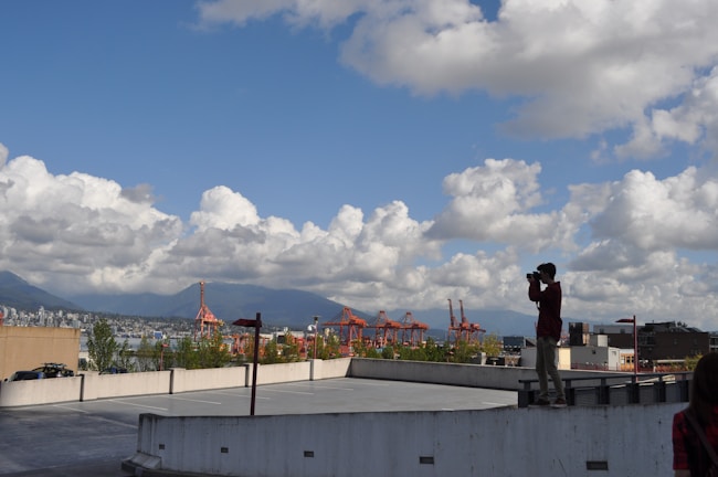 A person is standing on the edge of a rooftop parking area, holding a camera to take photographs. The sky is filled with fluffy white clouds, and in the background, there are red cranes of a port and a distant view of a city skyline with mountains.