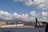 A person is standing on the edge of a rooftop parking area, holding a camera to take photographs. The sky is filled with fluffy white clouds, and in the background, there are red cranes of a port and a distant view of a city skyline with mountains.