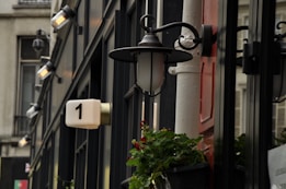 A close-up shot of a building facade features a series of wall-mounted lamps that emit a warm glow. A modern, rectangular plaque with the number '1' is visible, and a small pot of greenery adds a touch of nature to the scene. The facade is characterized by windows reflecting the adjacent building.