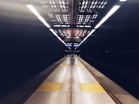 A subway station platform is depicted, showcasing an empty and symmetrical view with the focal point at the center. Bright overhead lights reflect off the dark polished surfaces, while yellow tactile paving marks the edge of the platform.