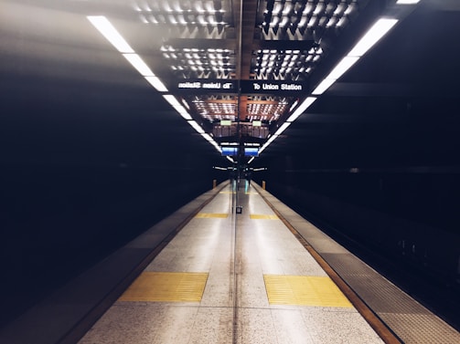 A subway station platform is depicted, showcasing an empty and symmetrical view with the focal point at the center. Bright overhead lights reflect off the dark polished surfaces, while yellow tactile paving marks the edge of the platform.