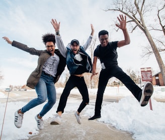 photo of three men jumping on ground near bare trees during daytime