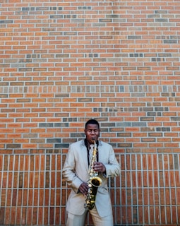 A person stands against a brick wall playing a saxophone. The person is wearing a beige suit and has closed eyes, suggesting concentration or enjoyment in the music. The wall consists of orange and gray bricks, providing a textured backdrop.