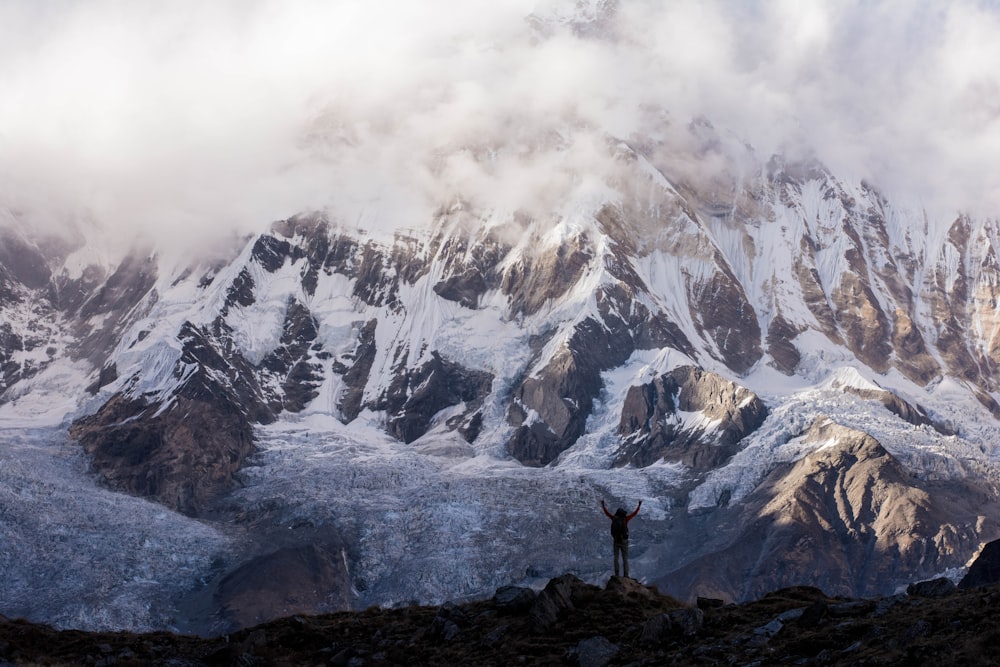 silhouette photo of a person raising hands on top of mountain near gray mountain covered with snow