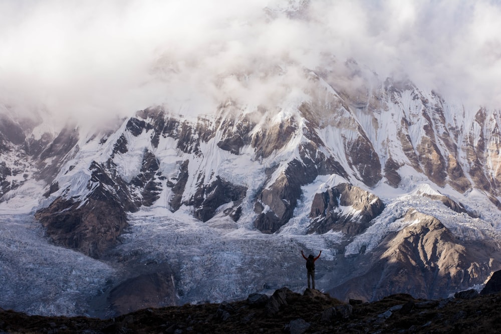 Silhouettenfoto einer Person, die die Hände auf dem Gipfel eines Berges in der Nähe eines grauen Berges hebt, der mit Schnee bedeckt ist