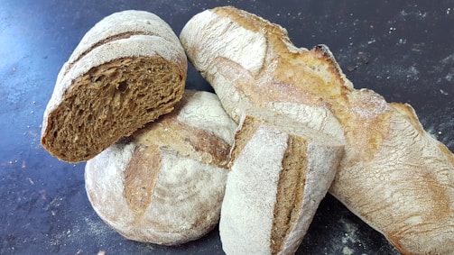 Several loaves of artisanal bread are scattered on a dark surface. The bread has a rustic appearance with a coated flour crust and varying shades of brown. One loaf is sliced open, revealing a dense, textured crumb.