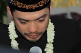 A man wearing a traditional black cap with intricate gold embroidery is captured in a close-up. He has tears in his eyes and is adorned with a garland of white flowers around his neck. His expression seems deeply emotional while he speaks into a microphone.