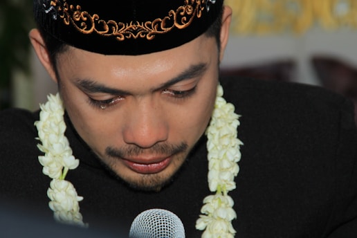 A man wearing a traditional black cap with intricate gold embroidery is captured in a close-up. He has tears in his eyes and is adorned with a garland of white flowers around his neck. His expression seems deeply emotional while he speaks into a microphone.