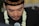 A man wearing a traditional black cap with intricate gold embroidery is captured in a close-up. He has tears in his eyes and is adorned with a garland of white flowers around his neck. His expression seems deeply emotional while he speaks into a microphone.