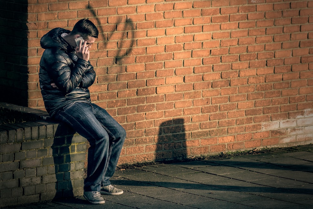 man in black leather jacket sitting on brick wall holding smartphone